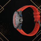 Right side profile of XTRI chronograph watch displaying matte dark gunmetal case thickness, crown, pushers, and red rubber strap on black and gold geometric backdrop.